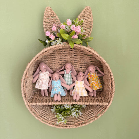 Five handmade fabric bunny dolls in floral dresses sitting in a woven bunny-shaped wall basket