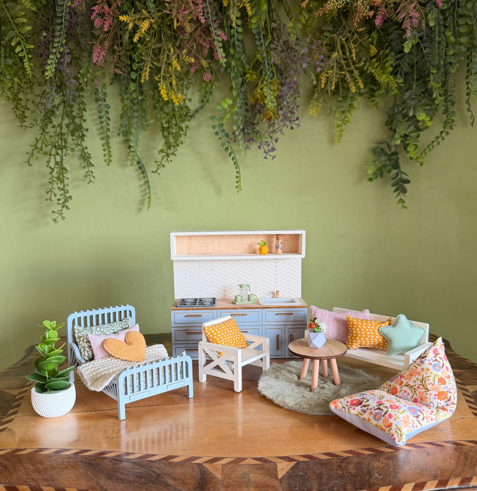 A complete view of a dollhouse furniture set on an antique wooden table in front of a green wall. The scene is fully furnished with a light blue kitchen and accessories, a white sofa and armchairs with pastel cushions, a small round table, and a light blue bed with a blanket and a yellow heart-shaped pillow.