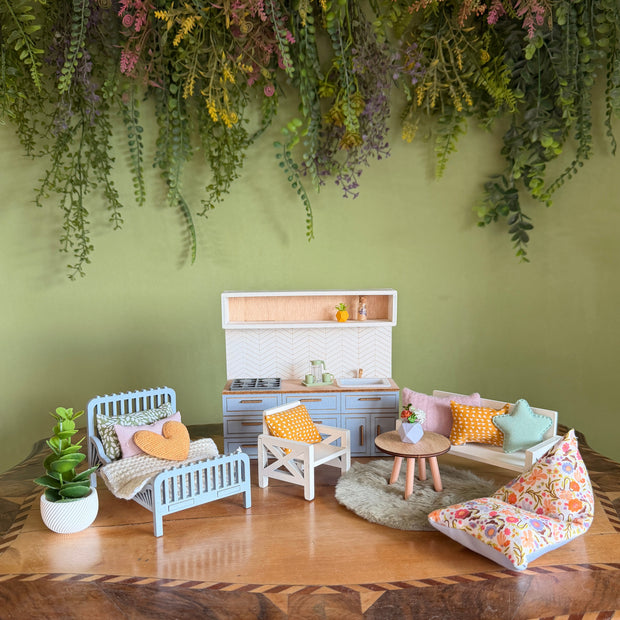 A complete view of a dollhouse furniture set on an antique wooden table in front of a green wall. The scene is fully furnished with a light blue kitchen and accessories, a white sofa and armchairs with pastel cushions, a small round table, and a light blue bed with a blanket and a yellow heart-shaped pillow.